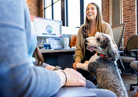 Over-the-shoulder view of a woman sitting at her desk conversing with a coworker while a dog stands with its paws in her lap